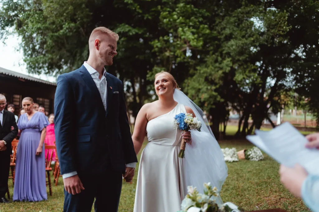 Ana e Fred sorriem na cerimônia no jardim, com as madrinhas de vestidos azuis e lilás