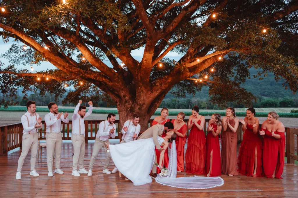 Bruna e Douglas com os padrinhos e as madrinhas embaixo de uma árvore iluminada, no fim da tarde