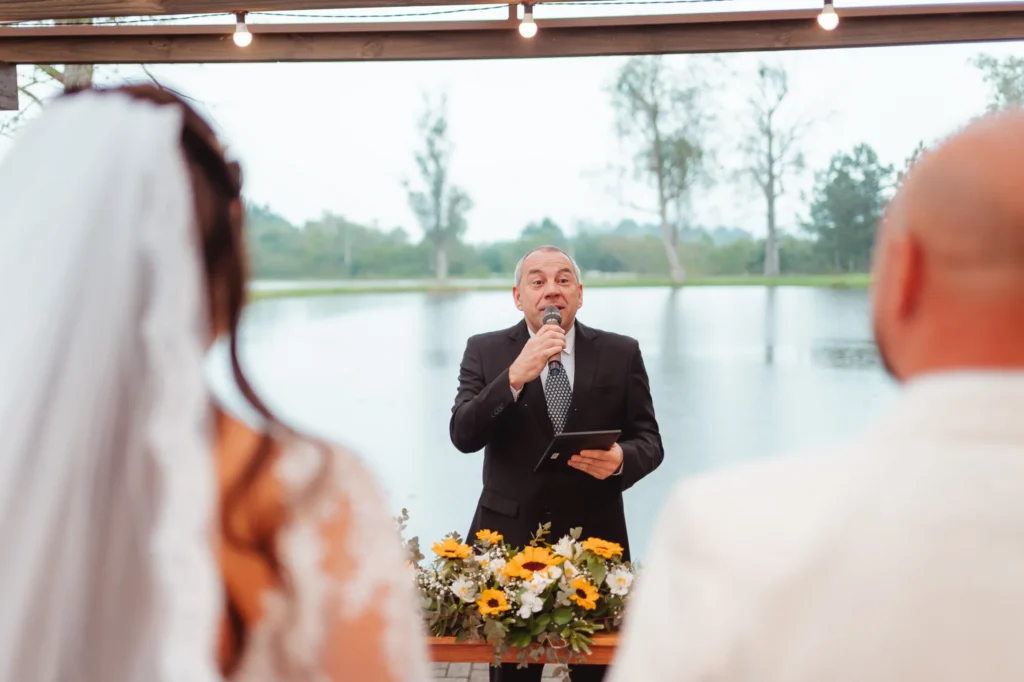 Celebrante fala ao microfone na cerimônia de Carla e Dionei, à beira do lago